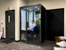 Load image into Gallery viewer, Person working in a black phone booth. Soundproof office pod for private phone call and distraction minimization. LED lights and ventilation for comfortable working sessions.